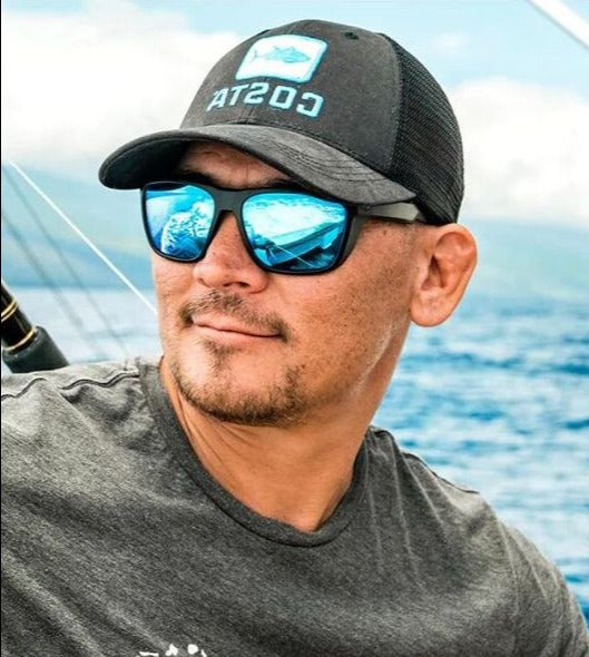 Man wearing reflective blue sunglasses and a black Costa hat, sitting on a boat with a fishing rod and ocean in the background.
