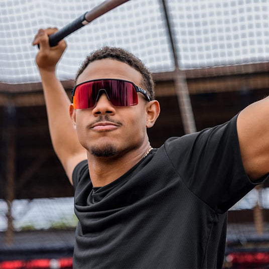 Man wearing red-lensed sports Oakley sunglasses and holding a baseball bat over his shoulders, standing in a baseball field.