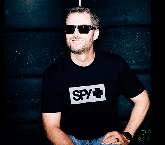 Dale-Earnhardt-Jr wearing spy optic sunglasses and wearing the brand t-shirt in front of a black wall