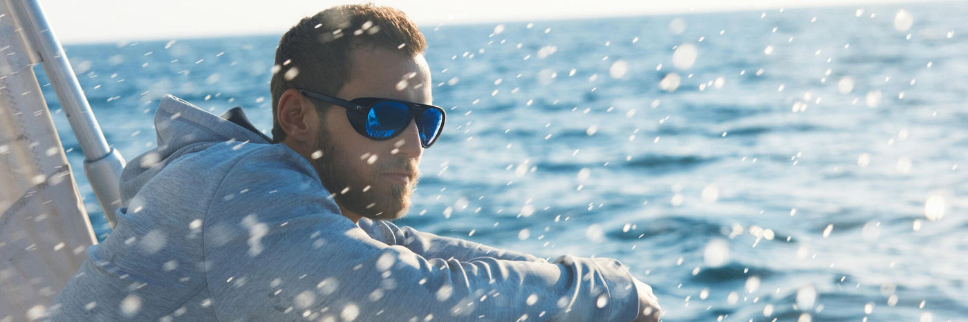 man on boat in middle of ocean wearing blue mirror costa del mar sunglasses while sea sprays into face